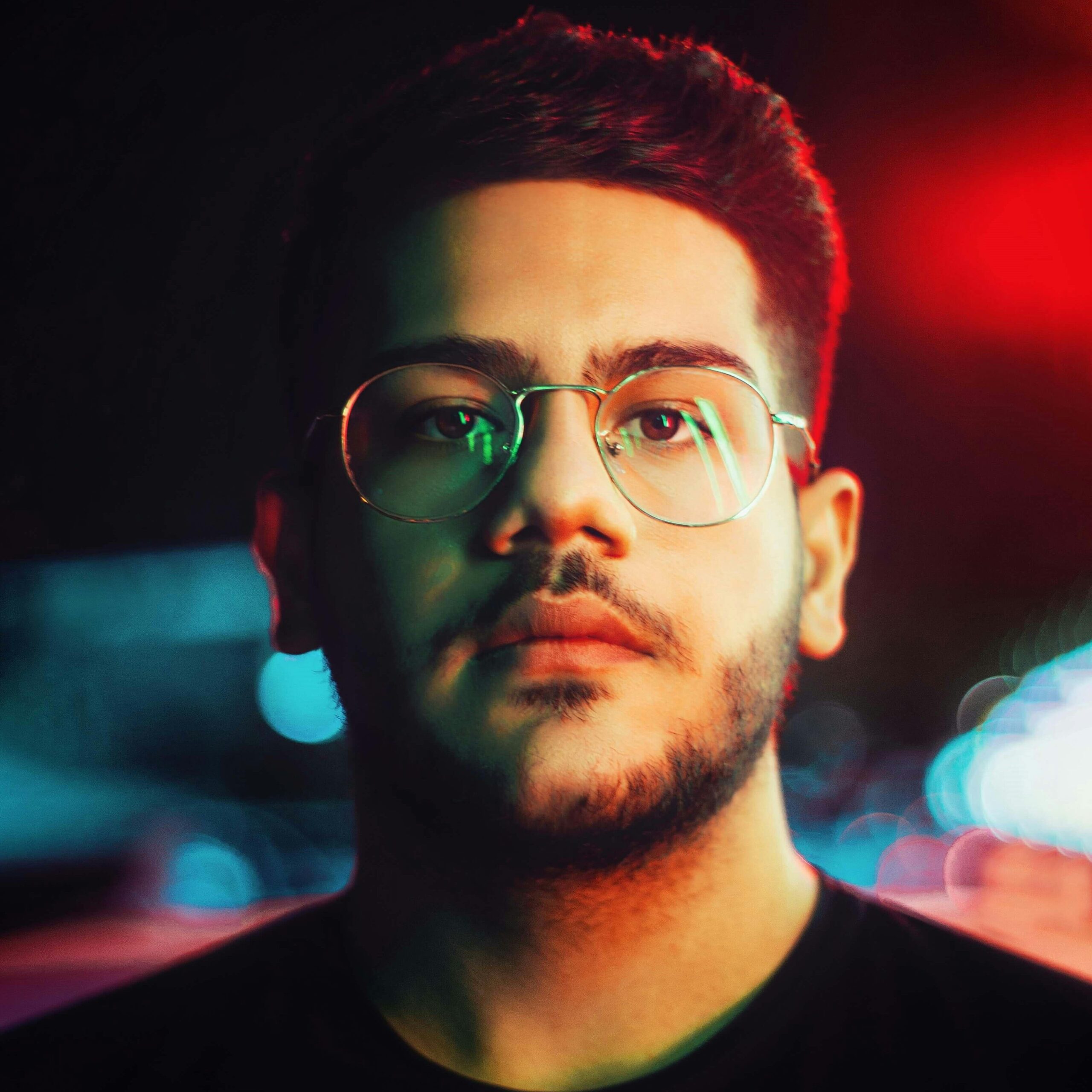 Vibrant night portrait of a young man with glasses and beard under colored lights.
