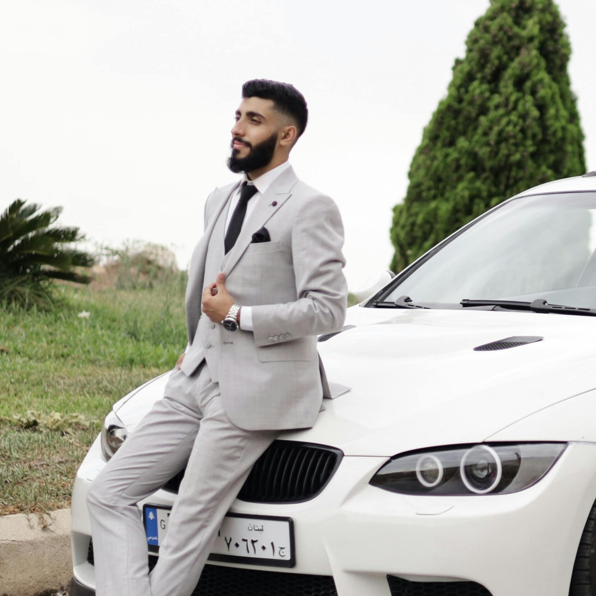 Elegant man in a suit leans on a white sports car, set against a scenic outdoor backdrop.